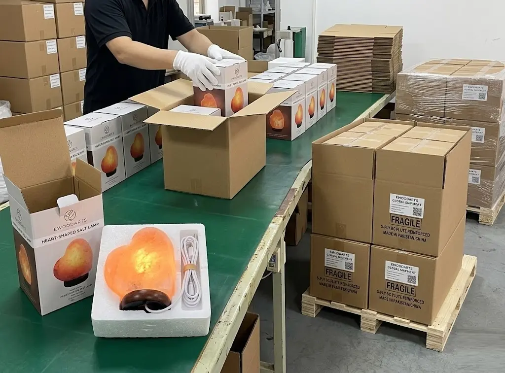 Professional packaging process for Himalayan salt lamps at Ewoodarts factory, featuring individual foam-protected heart-shaped lamps, custom color boxes, and reinforced 5-ply BC flute export master cartons on wooden pallets.
