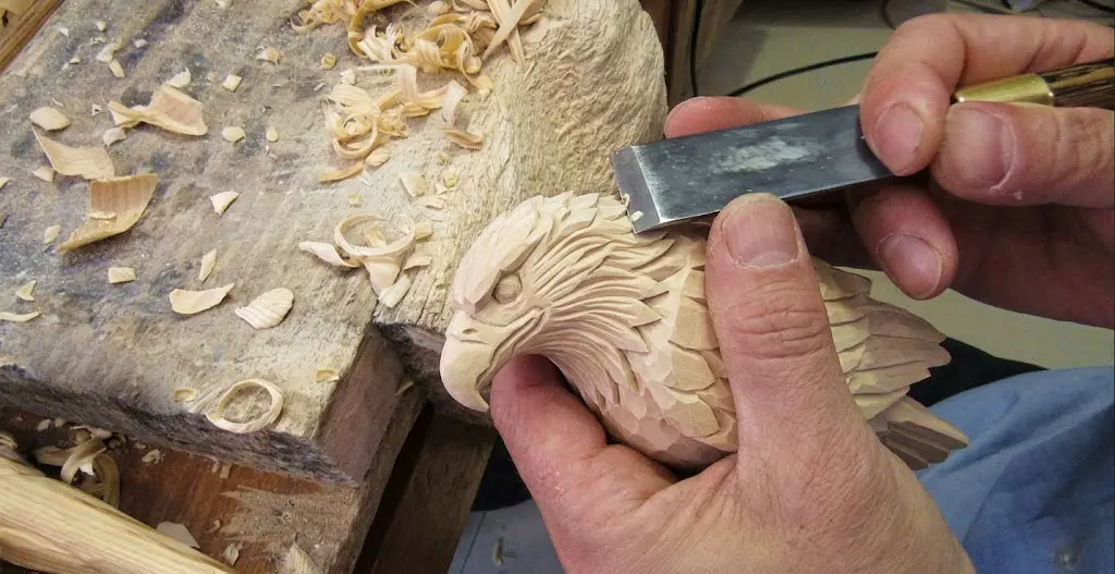 A macro photographic close-up of an artisan's hands meticulously carving an unfinished wooden eagle head from premium basswood. The left hand firmly holds the raw wood sculpture, while the right hand uses a sharp professional chisel to define the intricate plumage and sharp gaze. Realistic wood shavings and natural wood grain are scattered on the rustic workbench, capturing the 100% hand-carving process of ewoodarts.com.