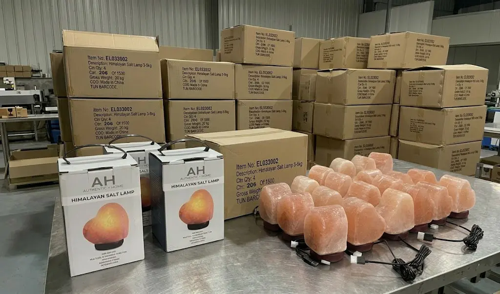 A professional factory scene showing a bulk order of heart-shaped Himalayan salt lamps on a stainless steel workstation, featuring custom 'AH' branded retail boxes and master cartons with detailed shipping marks and barcodes ready for export.