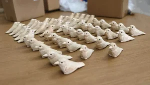 Sharp close-up of natural carved basswood birds with a blurred background showing a large production batch and wholesale boxes.
