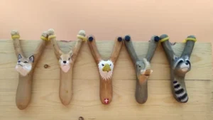 Hand-carved wooden animal slingshots including eagle, fox, and raccoon designs, showcasing detailed basswood craftsmanship for the wholesale gift market